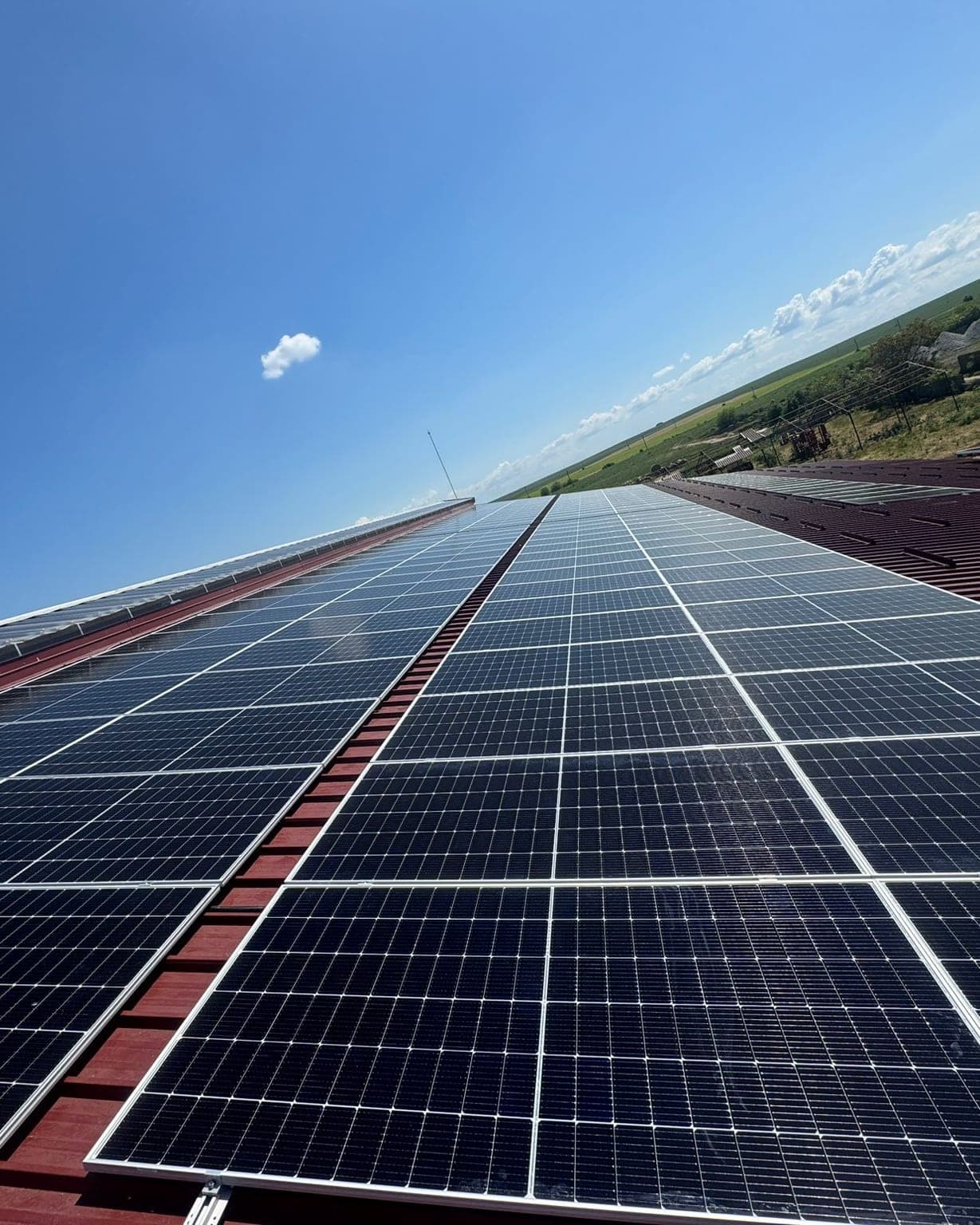 Solar panels along a pitched metal roof under a clear sky, with farmland beyond.
