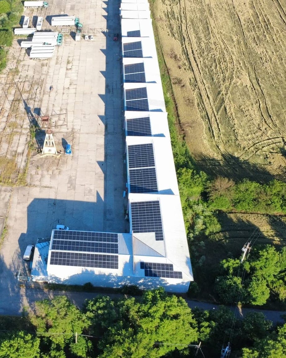 Aerial view of a long industrial hall with solar panels along its roof, beside fields and trees.